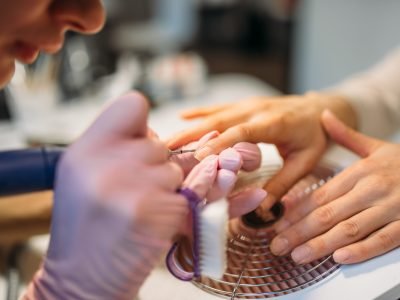 Beautician in gloves with polishing machine treats nails of female client, manicure in beauty salon. Manicurist doing hands care cosmetic procedure